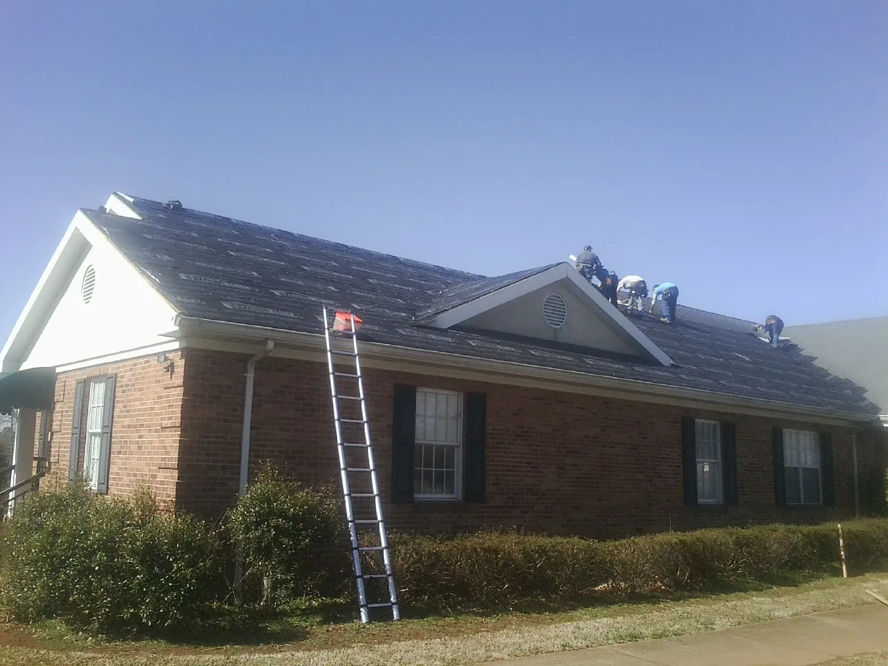 Crew tearing shingles off a brick ranch roof with a ladder set — Metal Roof Repair work by our crew in Dahinda