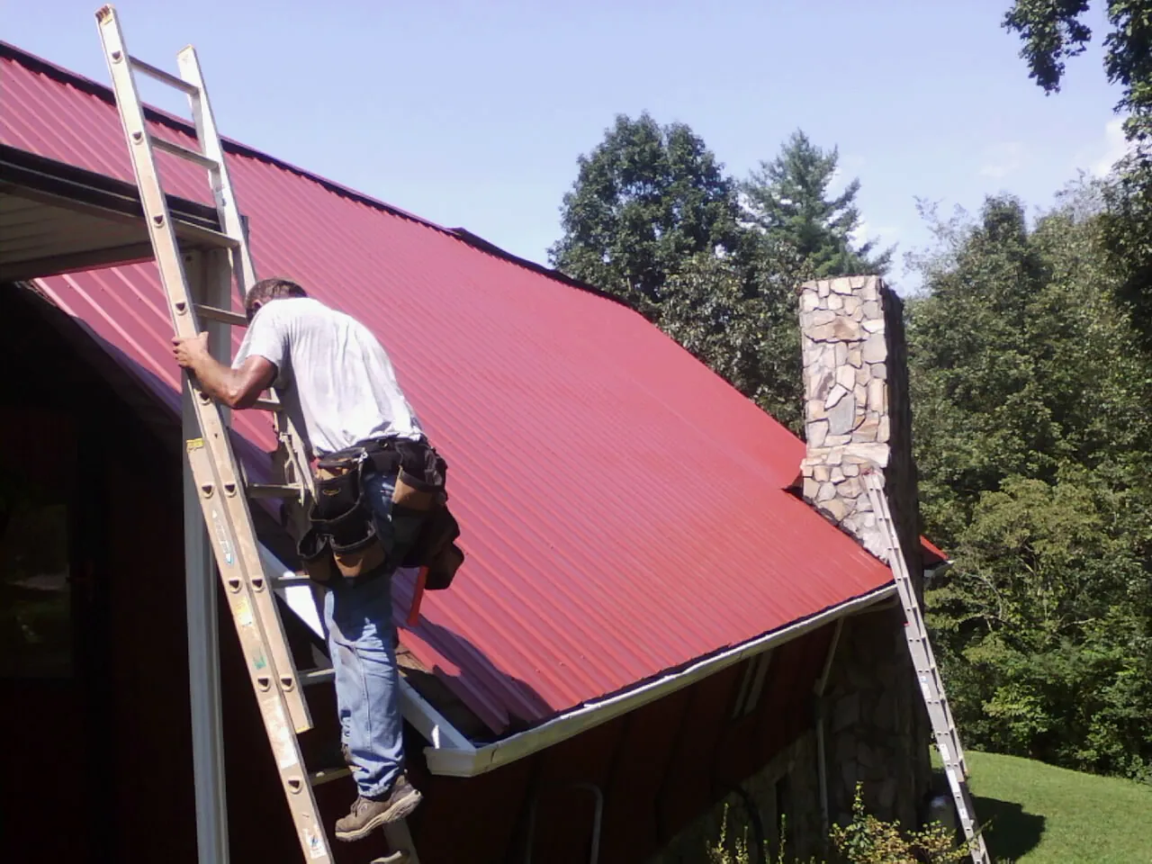 Roofer on a ladder beside a red metal roof and stone chimney — Soffit & Fascia Repair by Roofing Brothers in Dahinda